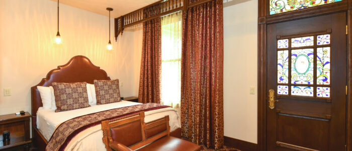 Room 10 Finch Guest House Queen Bed with Two Nightstands and 2 Pendant Reading Lights. Antique Eastlake One-Arm Ladies Chair at Foot of Bed. Spectacular Door to Front Porch Stained Glass with Stained Glass Transom. Victorian Fretwork Detail Length of Ceiling and Heavy Burgundy and Gold Drapery