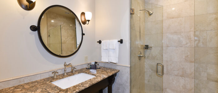 Room 11 Bathroom Features Glass Enclosed No-Curb Shower with Large Porcelain Tile. The Granite Topped Vanity Has Sink and Wall-Mounted Tiltable Mirror.