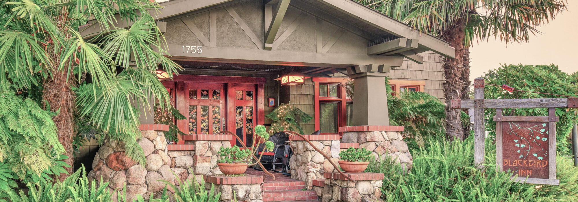 Craftsman Style Blackbird Inn rusty metal sign with turquoise ginko tree leaf design. Main color dark green shingle, mahogany door and window frames, River rock base around front porch and post bases. Palms on both sides of building, ferns against river rock.