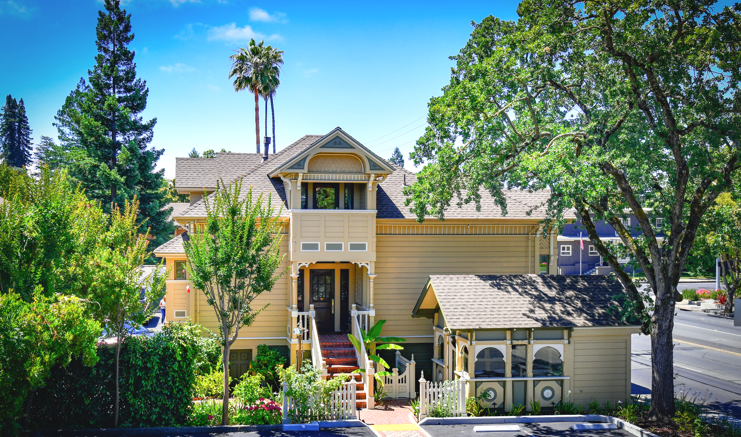 Eastlake, Queen Anne Styled Finch Guest House. Steep Red brick staircase leads to entry porch. Above Entry porch, a 2nd story cover porch mimics form of classic Victorian tower. Smaller building to right is gazebo. Landscaping is large valley oak and topicals near building.