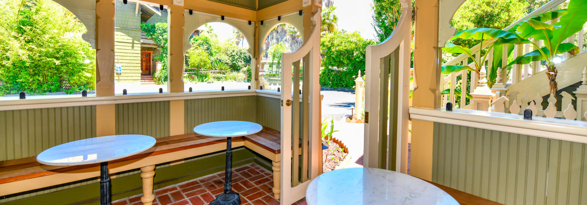 Interior of gazebo. Floor is red brick pavers, walls are Victorian styled wainscoting with arched openings pain colors are green, yellow and cream. 3 marble topped tables are in front of built-in wooden benches. Outside windows are plantings and bananas.