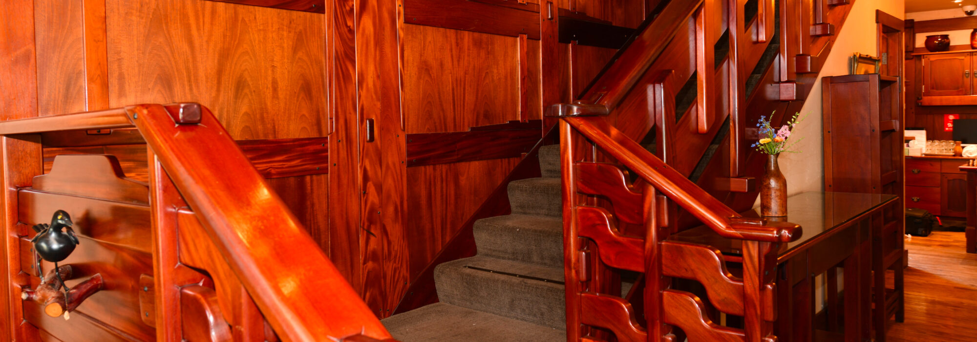 Main staircase of Blackbird Inn. Carved mahogany balustrade in wavy craftsman pattern surrounds green carpeted stairs. Back wall is mahogany paneling.
