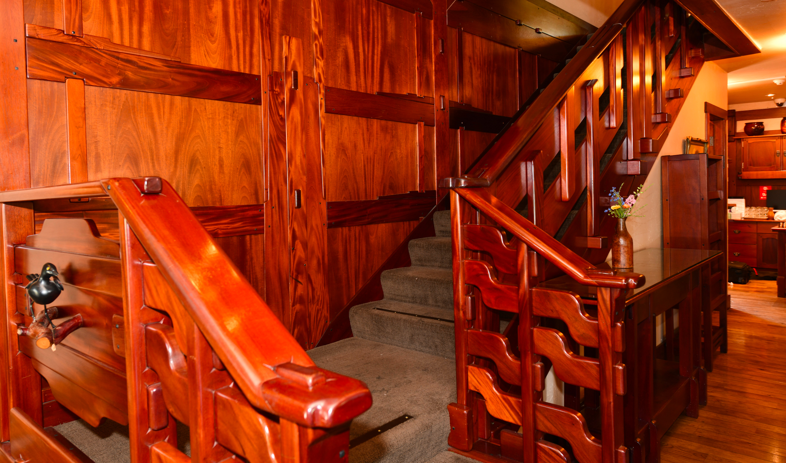Main staircase of Blackbird Inn. Carved mahogany balustrade in wavy craftsman pattern surrounds green carpeted stairs. Back wall is mahogany paneling.